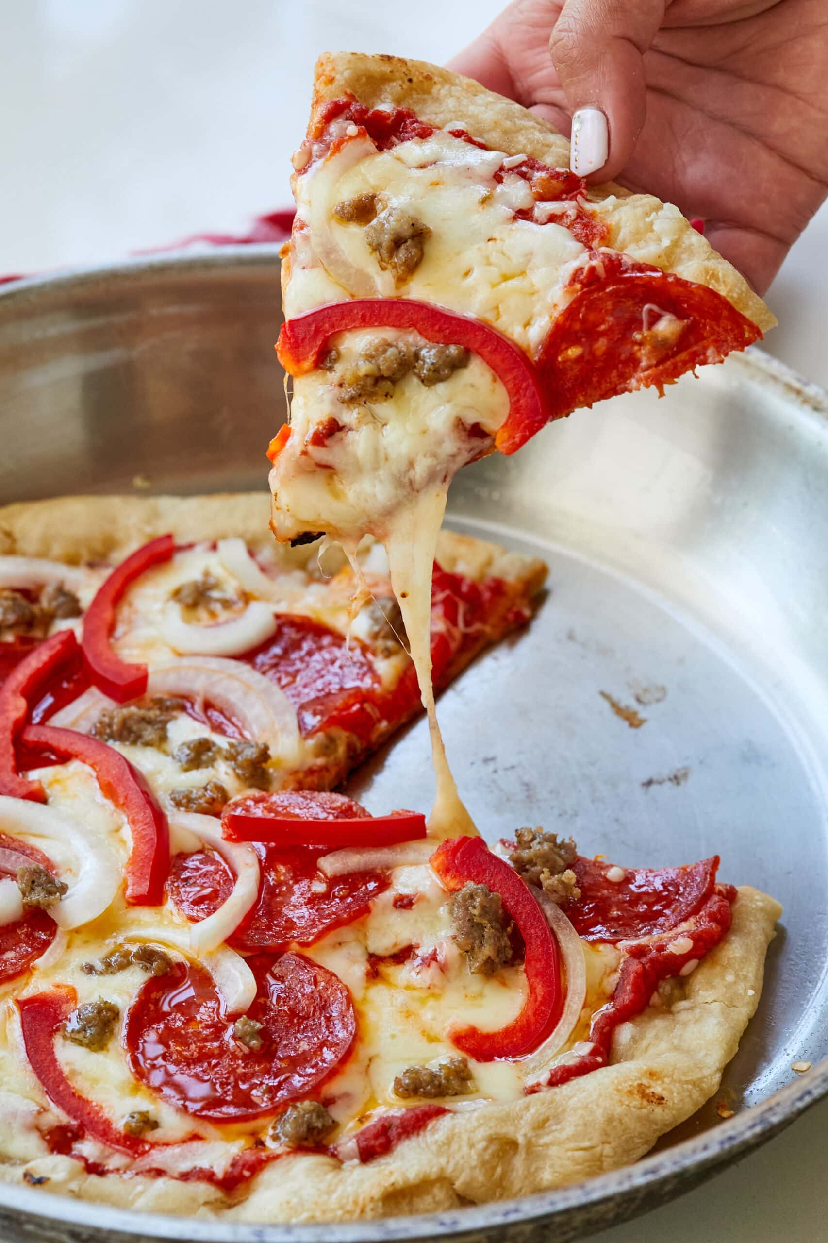 A slice of Pan-Fried Pizza is being lifted from the pan, with crispy edge, melted cheese, pepperoni and red bell paper strips.