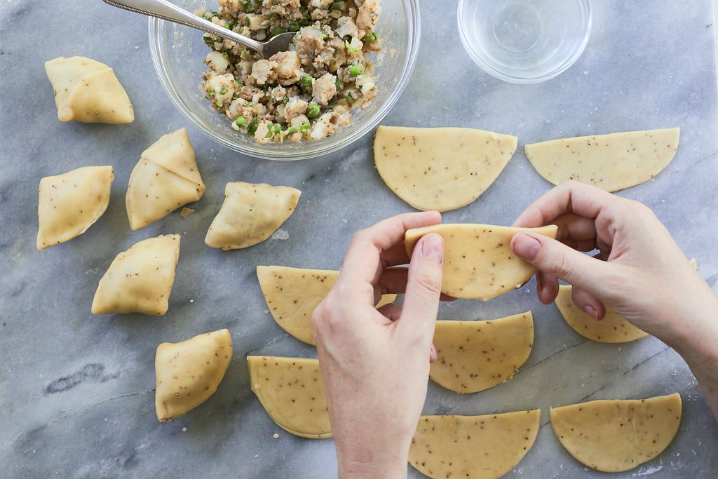 Step one of folding and filling a homemade samosa, with dough ready to use and being held by hands and filling to be spooned.