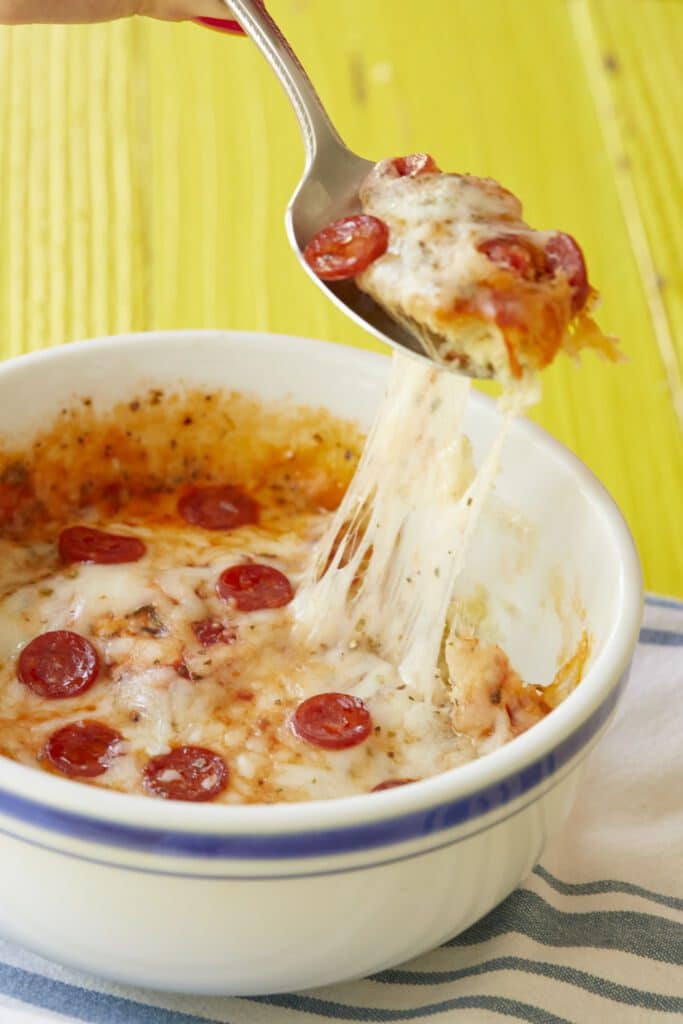 A scoop of pizza is being lifted from the pizza bowl with melted mozzarella being stretched very long. It is lightly crispy on the edge along the bowl, has gooey cheese, caramelized pepperoni and aromatic dried herbs on top.