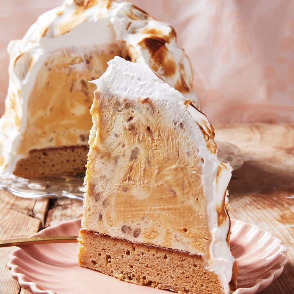 A slice of Banoffee Baked Alaska is served on a dessert plate. It has a blondie base topped with a mixture of caramel ice cream, banana ice cream, and salted caramel sauce, topped with meringue and toasted until golden brown.
