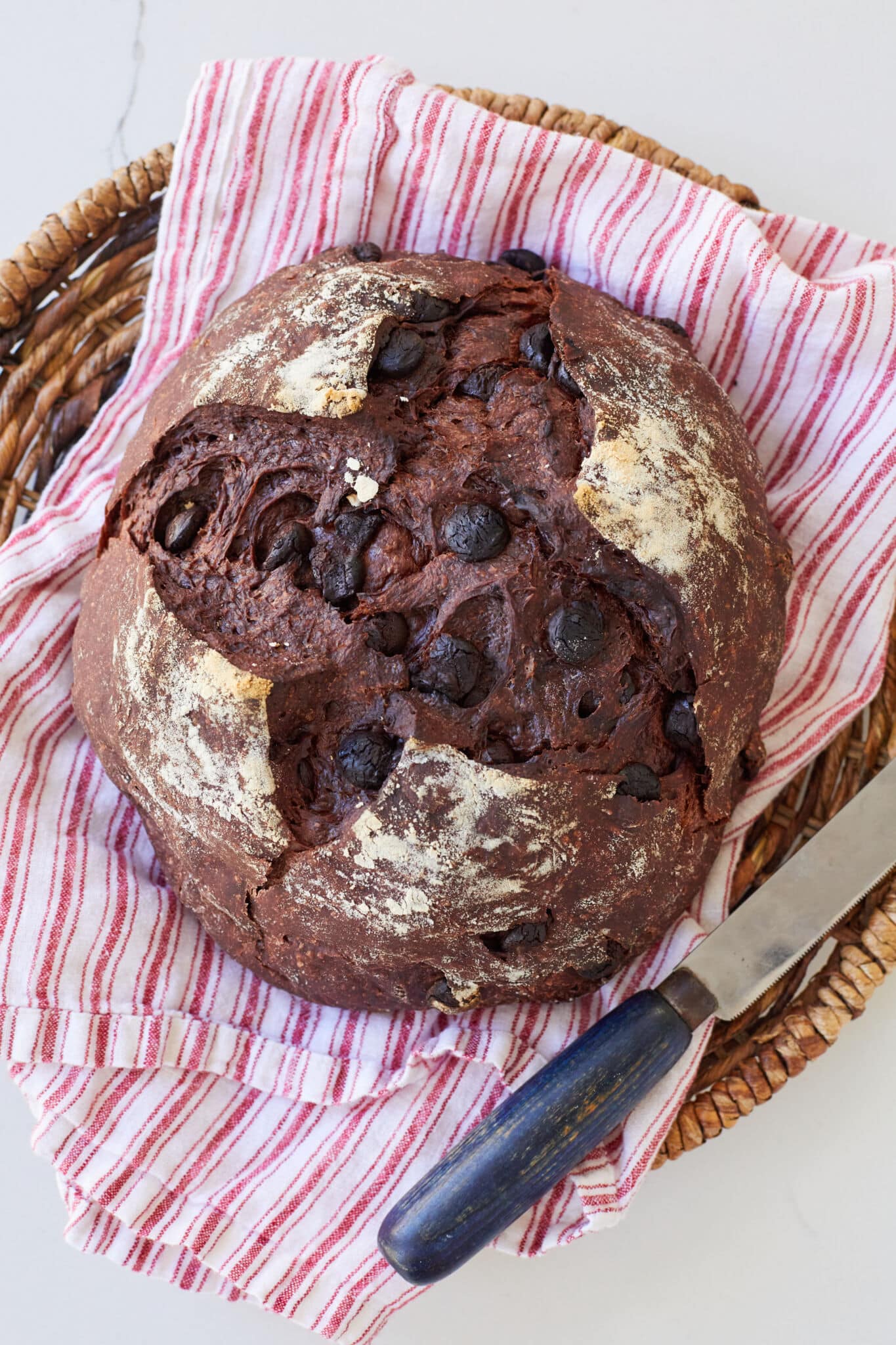 A loaf of Chocolate Bread is on a kitchen towel in a bread basket with a bread knife. It's a dark-chocolate color loaf with crispy crust and a cross score on top, loaded with chocolate chips. The inner crumb looks very bubbly.