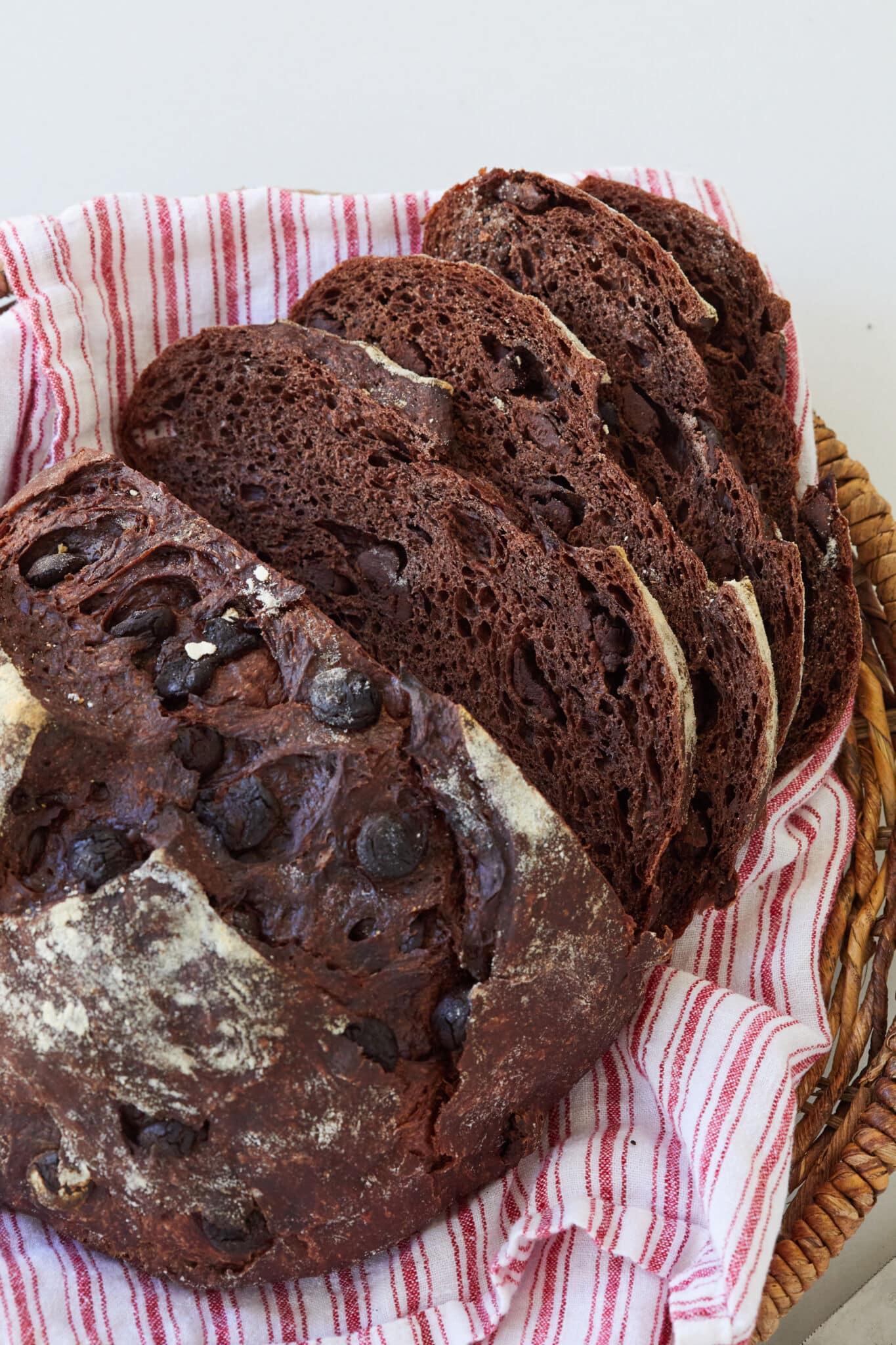 A close-up shot at loaf of Chocolate Bread on a kitchen towel in a bread basket. It's sliced and has a dark-chocolate color with crispy crust and a cross score on top, loaded with chocolate chips. The inner crumb looks very tender and bubbly.