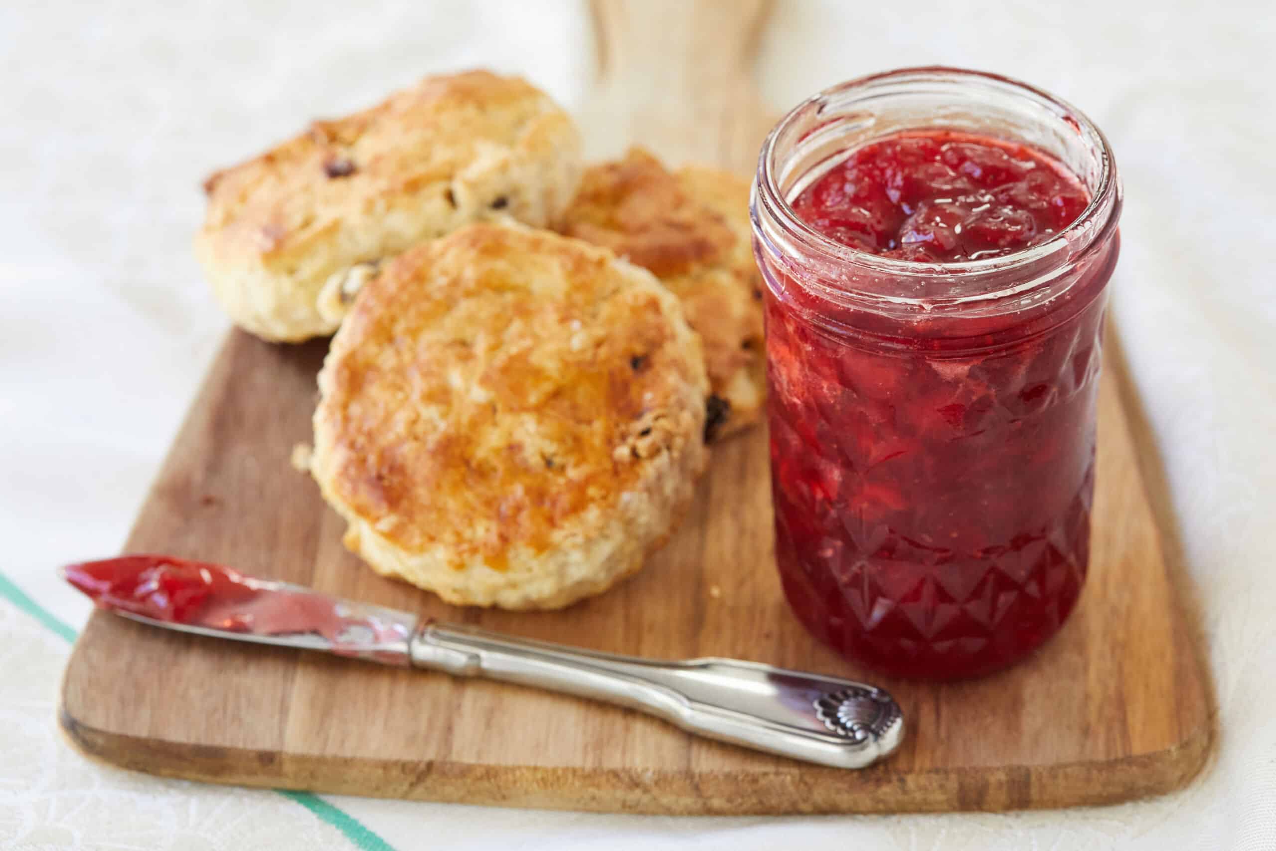 Vibrant red, thick Classic Strawberry jam is in a glass jar next to scones.