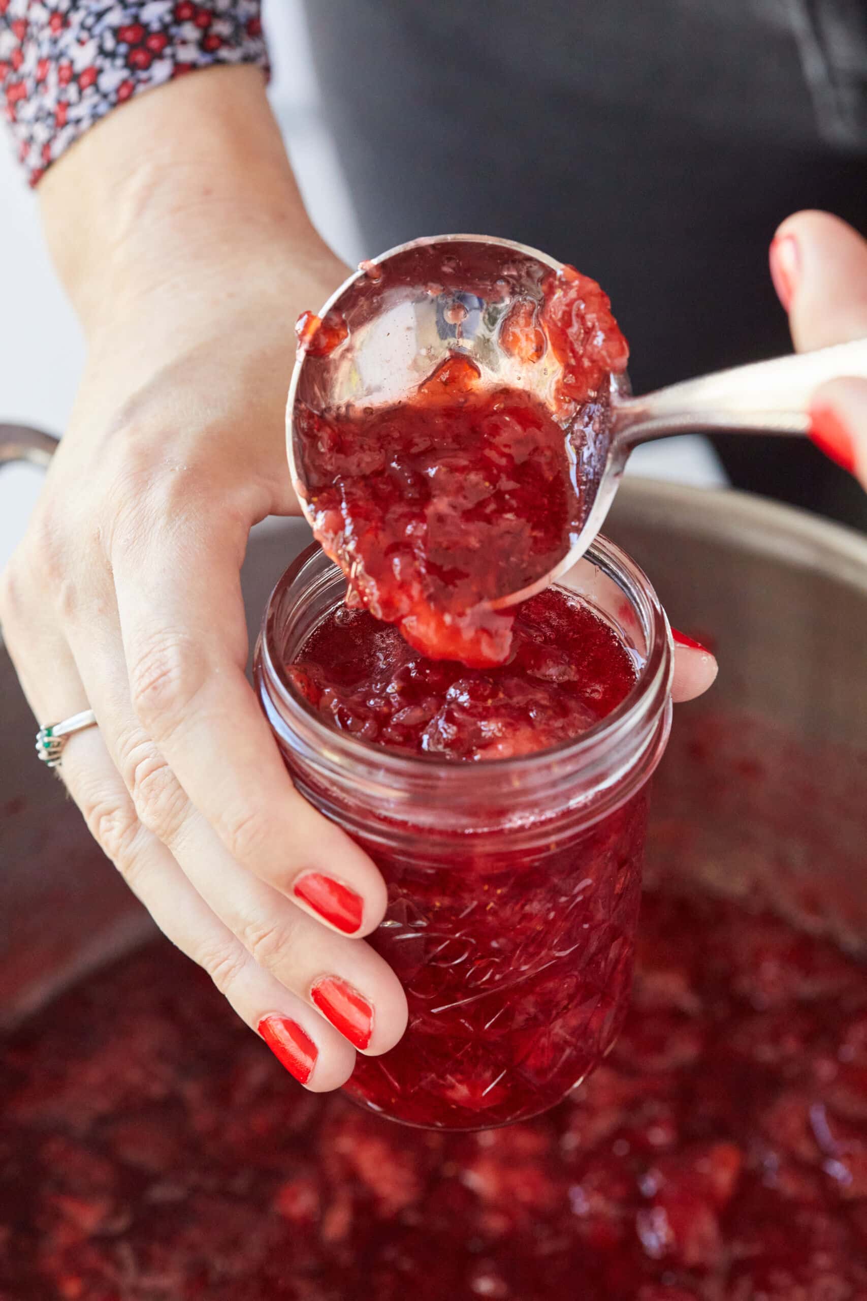 A spoonful of thick, vibrant-red Classic Strawberry Jam with Pectin above a glass jar over the big pot.