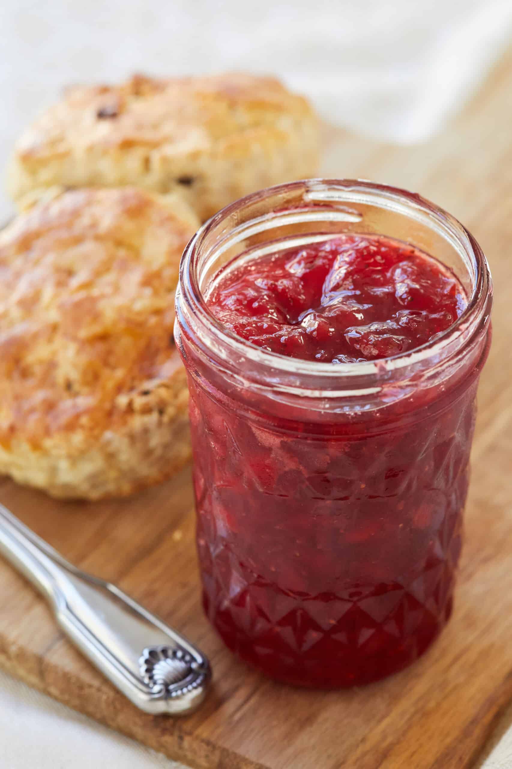 Vibrant red, thick Classic Strawberry jam is in a glass jar in front of two scones.