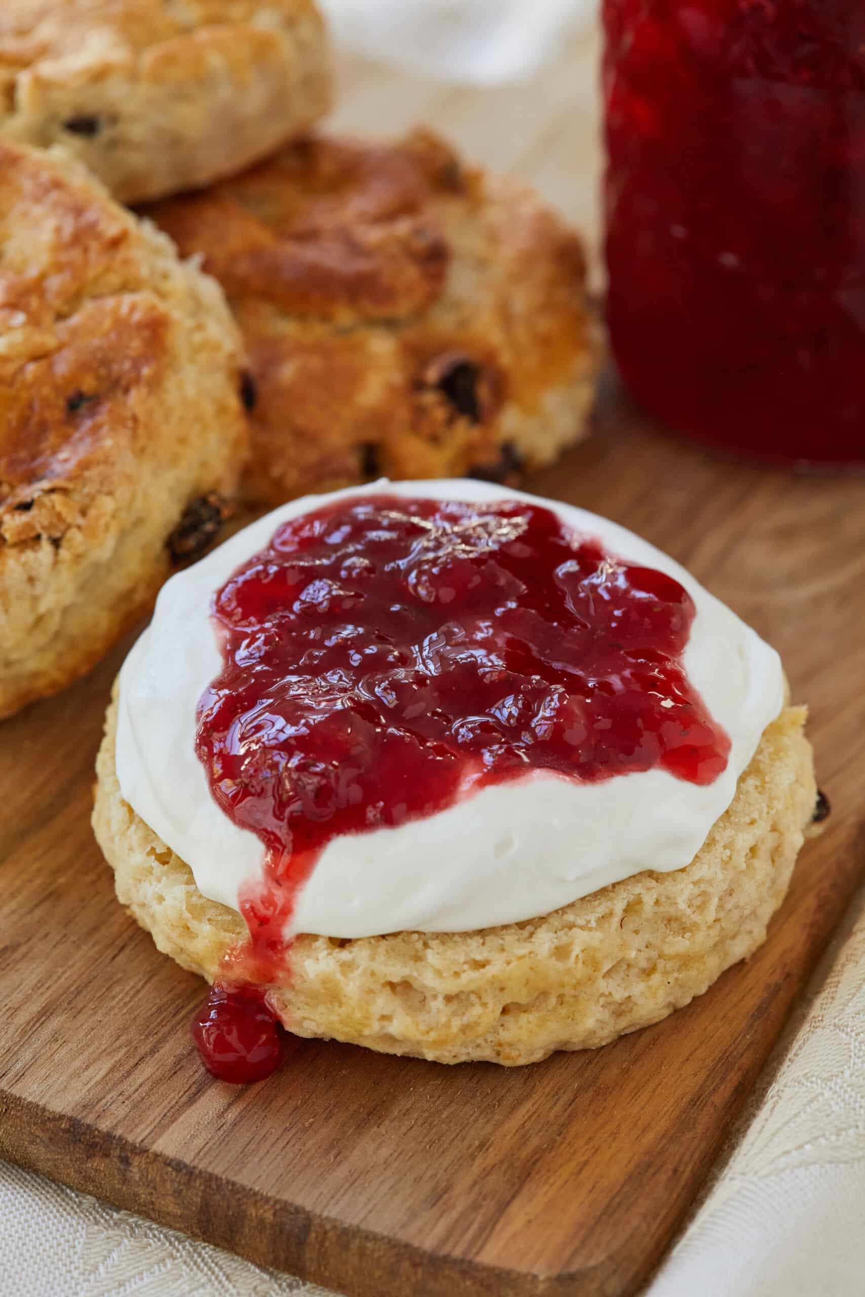 Vibrant red, thick Classic Strawberry jam is put on top of whipped cream to pair with scones. More scones are in the back ground.