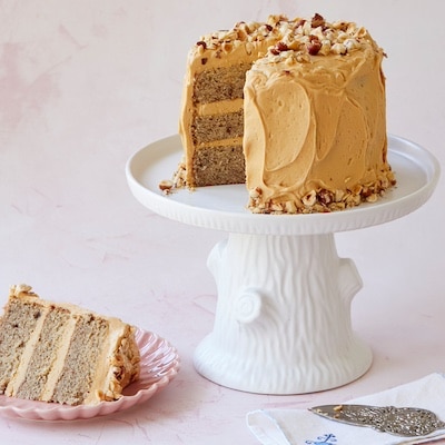A close-up shot at the cut of Hazelnut Cake shows its 3 layers of cake and 2 layers of filling, Salted Caramel Buttercream Frosting on the outside, and decorated with toasted nuts on top and around the bottom. A slice os served on a dessert plate next to the white cake stand.