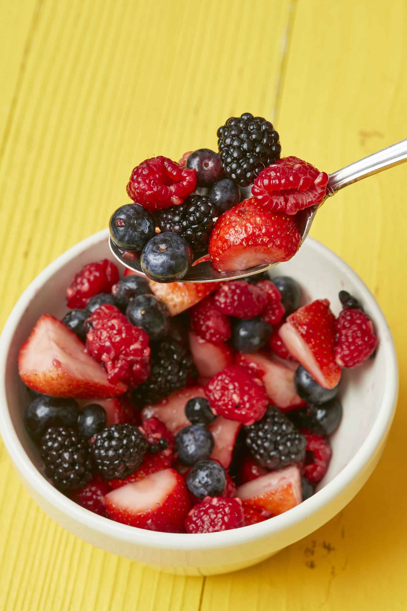 A bowl and spoonful of macerated fruit including strawberries, blueberries, blackberries, raspberries.