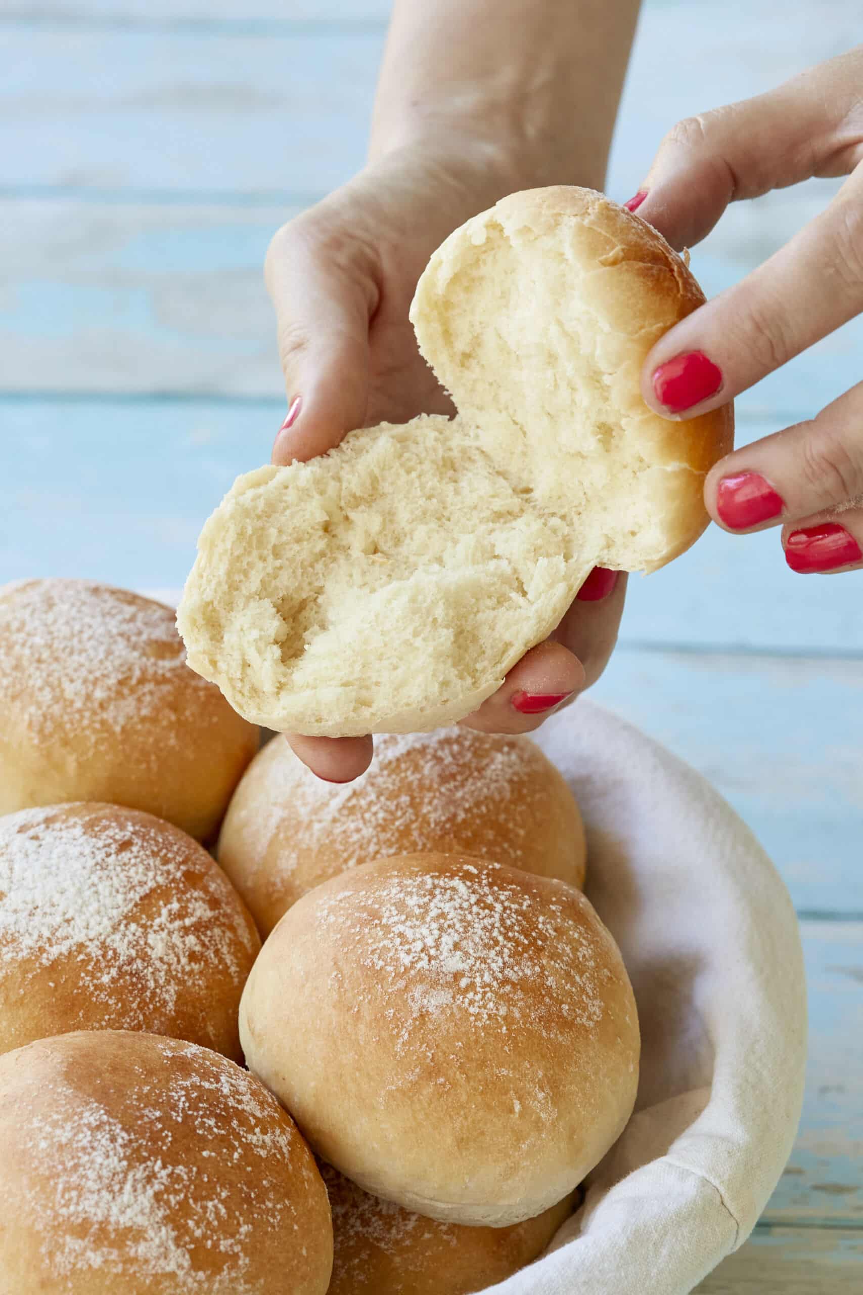 A basket of soft, fluffy Scottish Baps are lightly dusted with flour. One is sliced open showing the bully crumb.