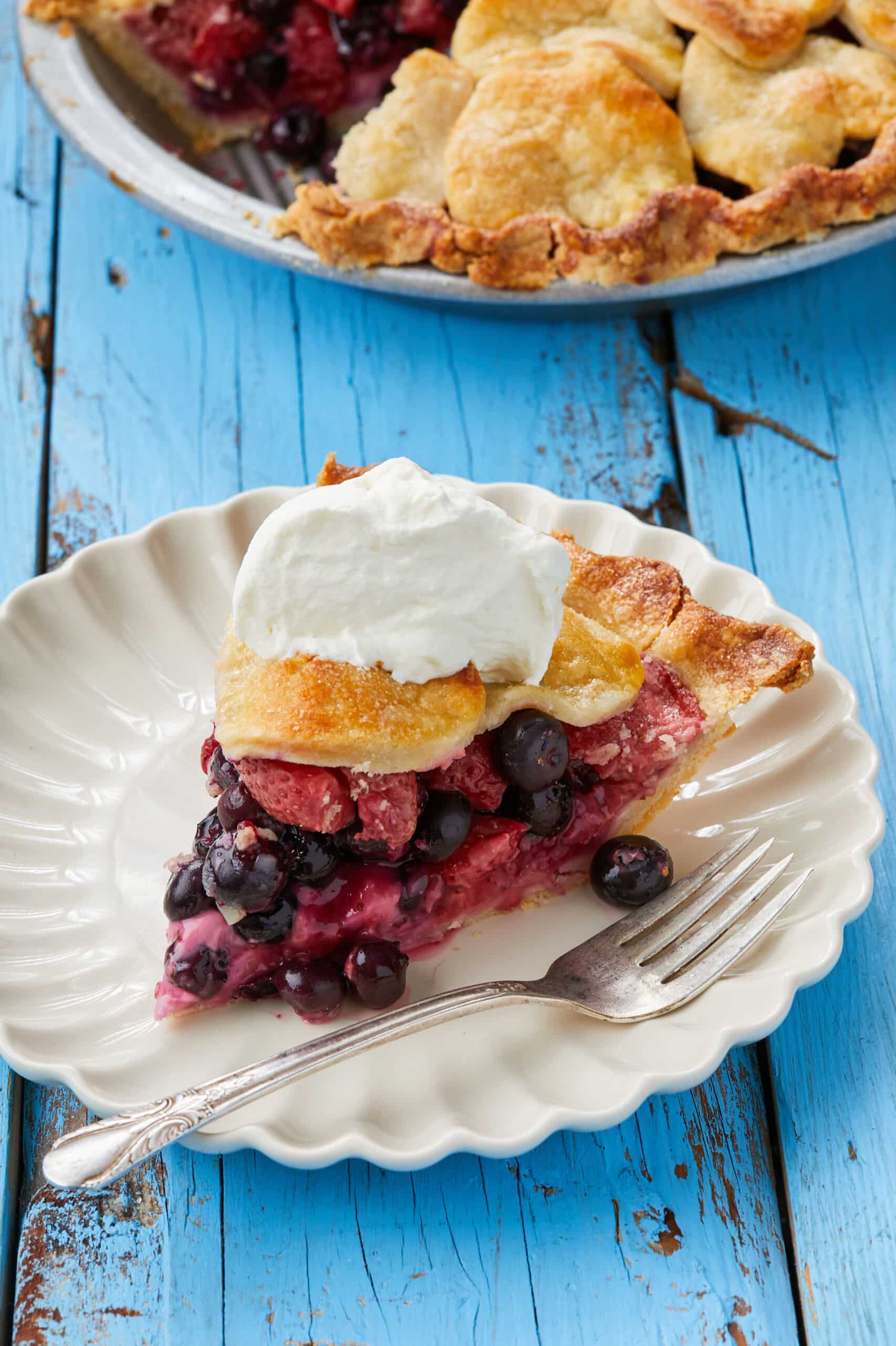 A close-up shot at a slice of the Blueberry Strawberry Pie shows its flaky crust, fruity juicy filling, served with a dollop of whipped cream.