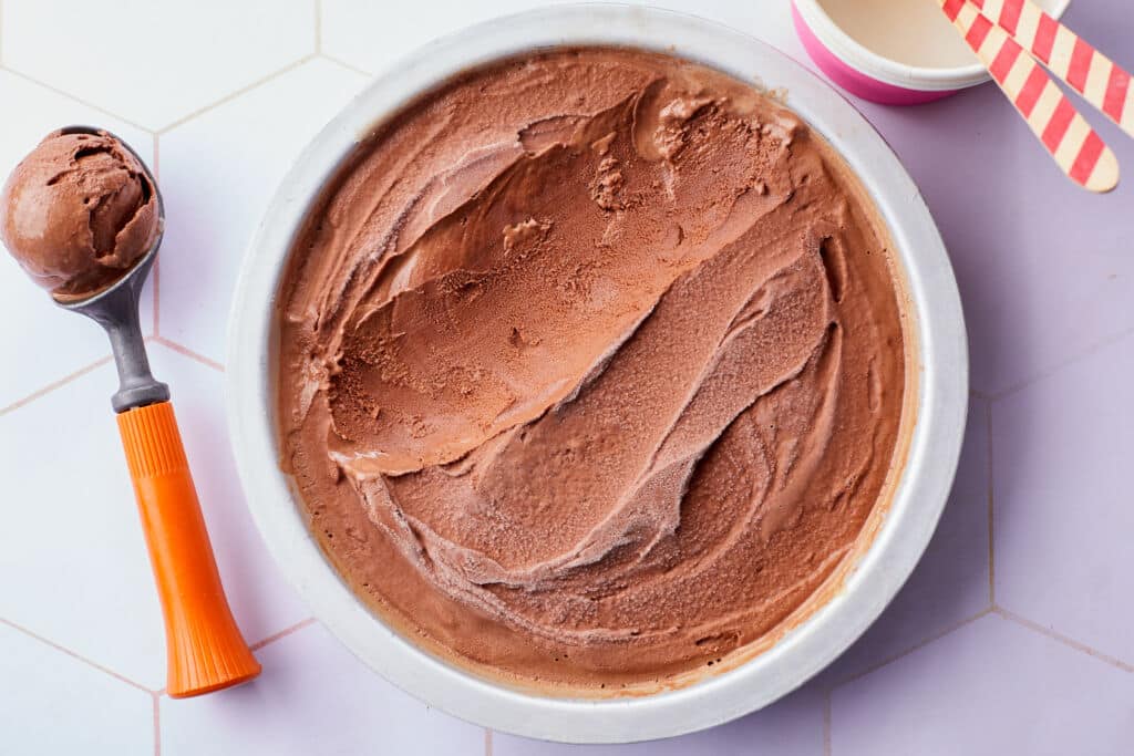 The smooth and thick Healthy Chocolate Frozen Yogurt is chilled and ready to serve. A coop of the frozen yogurt and ice cream cups with spoons are on the side.