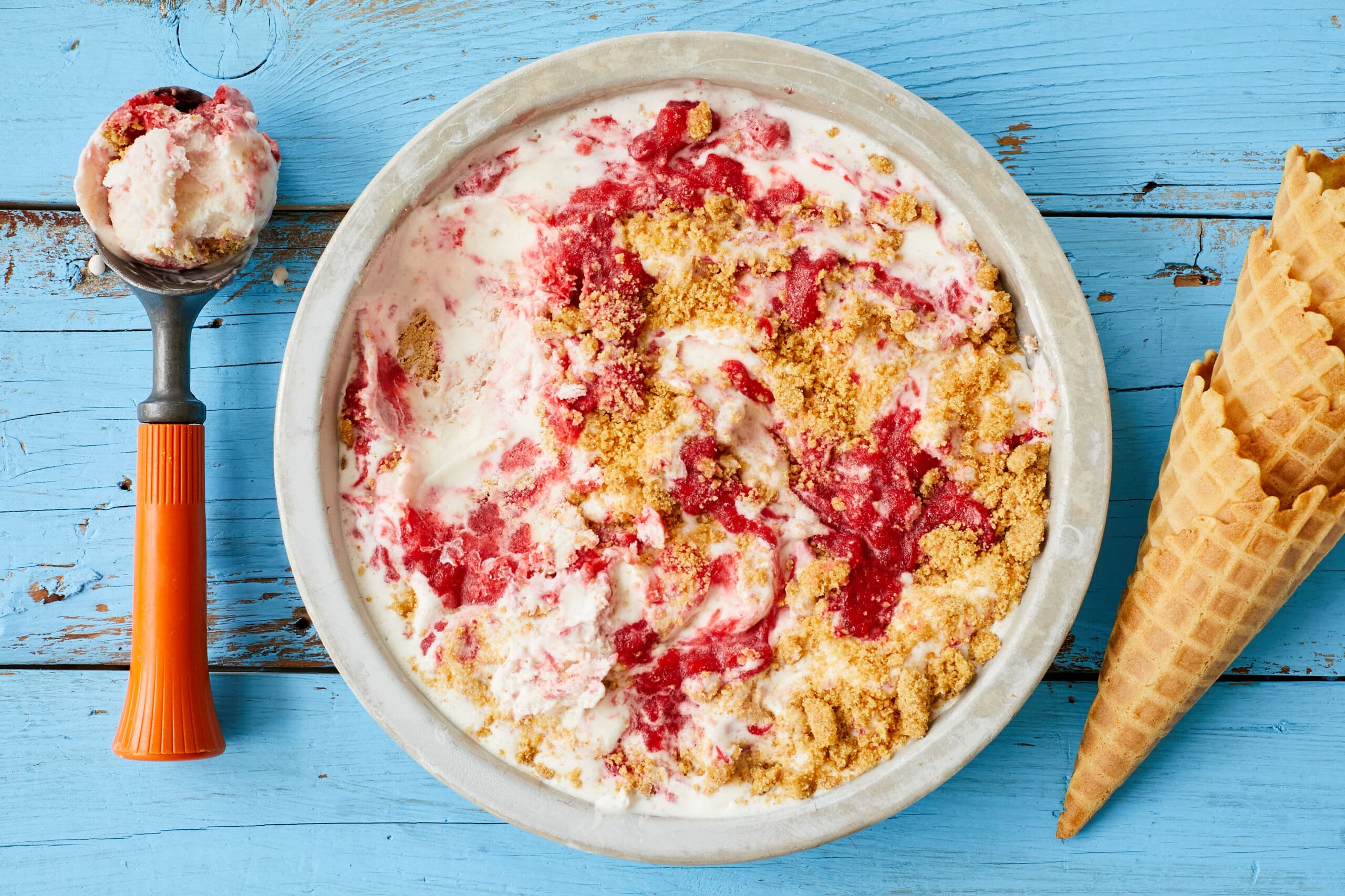 An over-head shot of a bowl of Strawberry Cheesecake Ice Cream shows the creamy, thick ice cream with swirls of jammy strawberries and chunks of buttery Graham cracker crust. An ice cream scoop is on the left side and an ice cream cone is on the right.