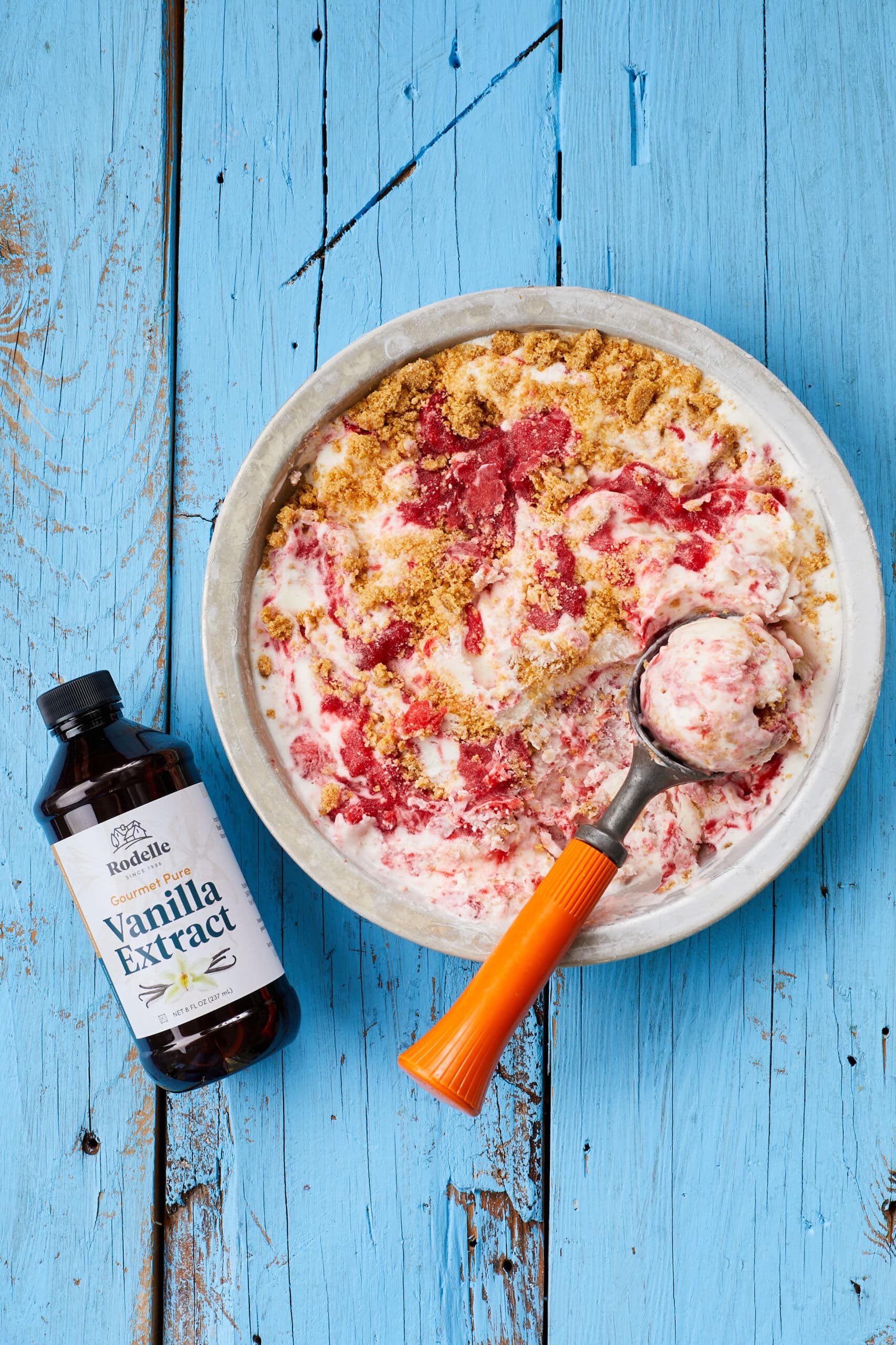 An over-head shot of a bowl of Strawberry Cheesecake Ice Cream shows the creamy, thick ice cream with swirls of jammy strawberries and chunks of buttery Graham cracker crust. A bottle of vanilla extract is on the side.