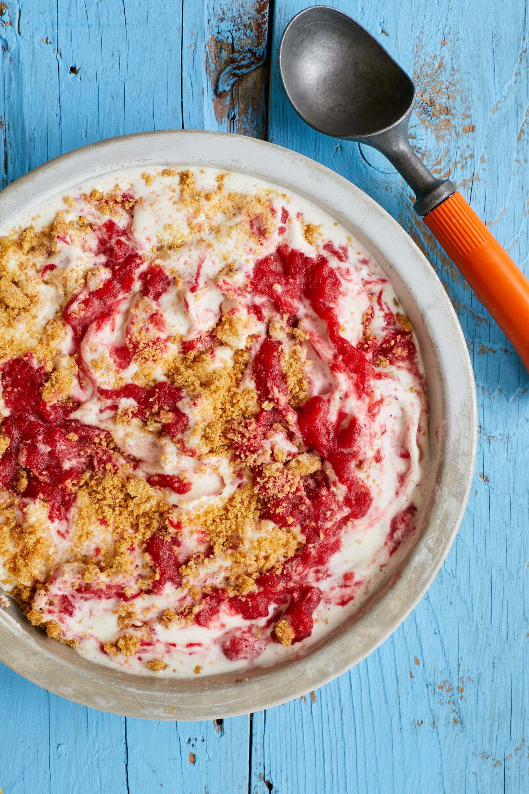 An over-head shot of a bowl of Strawberry Cheesecake Ice Cream shows the creamy, thick ice cream with swirls of jammy strawberries and chunks of buttery Graham cracker crust. An ice cream scoop is on the side.