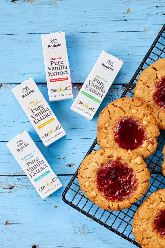 Peanut Butter and Jelly Thumbprint cookies are baked until golden brown with jelly in the centers and crackled tops. They're served perfectly with milk. Four Rodelle Single-Origin Pure Vanilla Extract are on the let side.