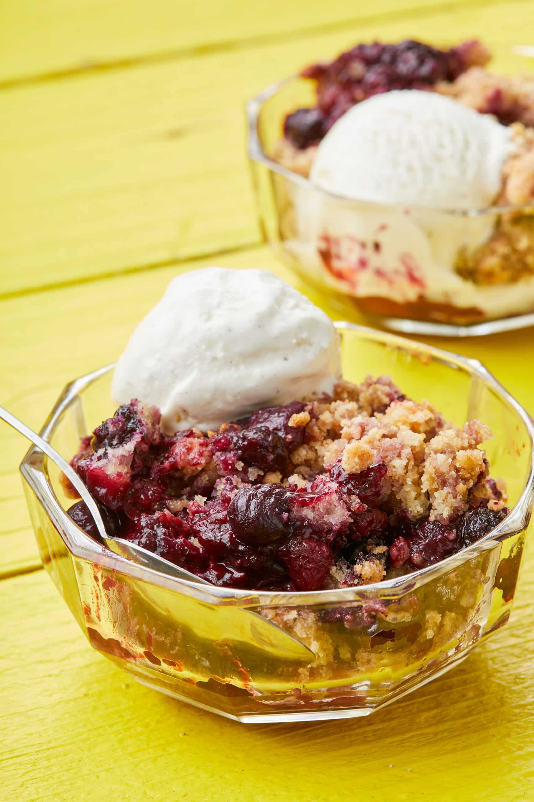 A close-up shot at Mixed Berry Cobbler with Cake Mix in a glass dessert bowl, with soft berries, golden cobbler, and vanilla ice cream on top.